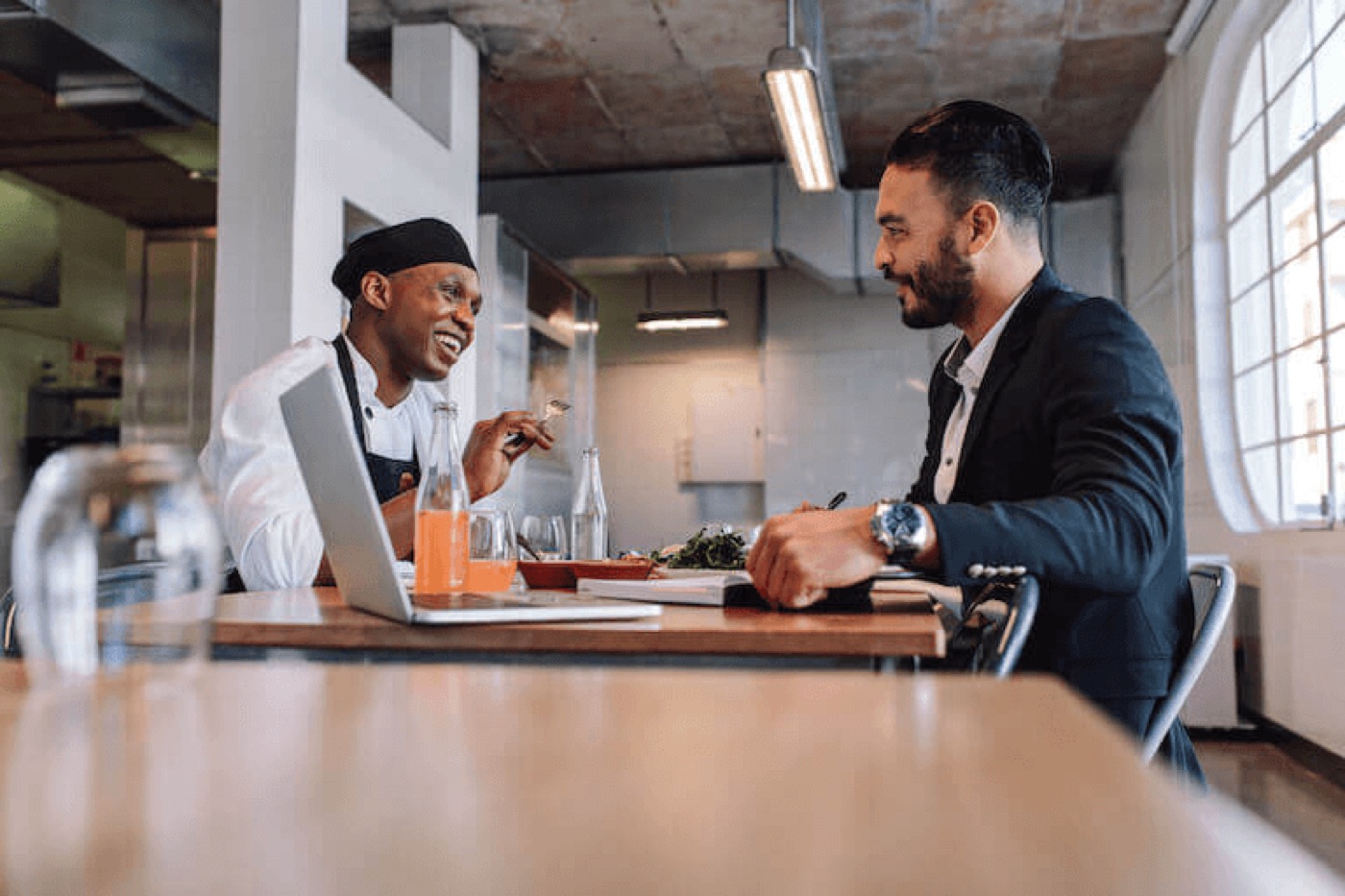 A chef and a business partner sitting across a table with a laptop, talking and smiling over drinks and a shared meal