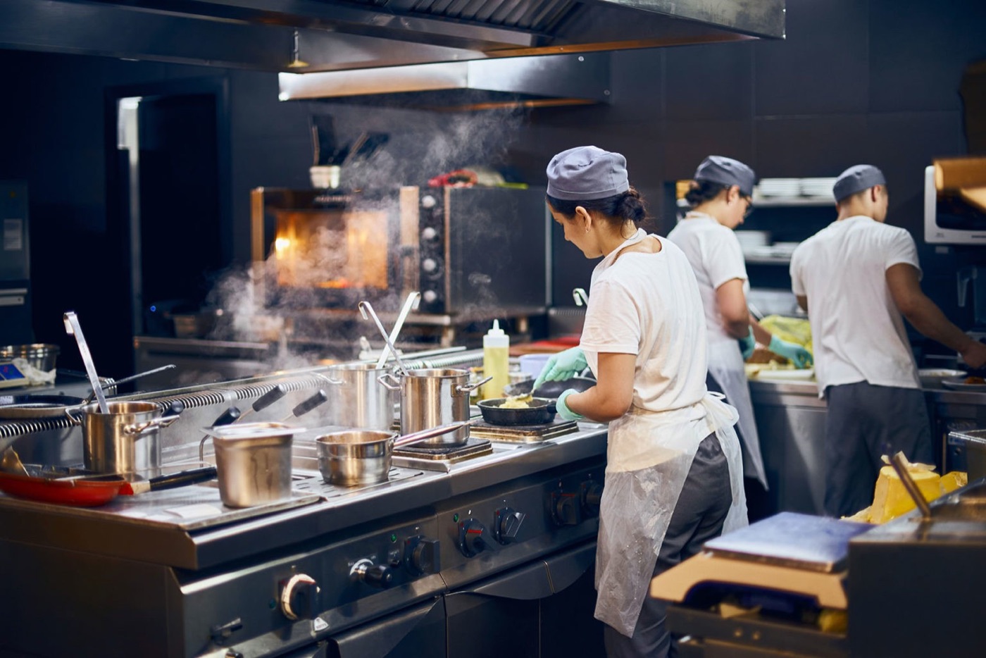 Kitchen staff in a commercial kitchen preparing food together at the stove, with pots steaming and an oven glowing in the background
