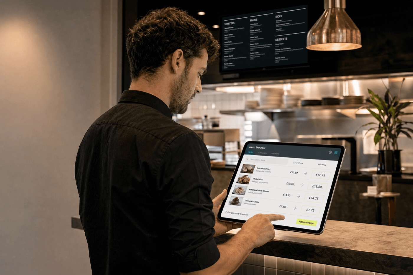 Restaurant staff member updating menu prices on a tablet using a menu management app, with a digital menu board visible in the kitchen behind them